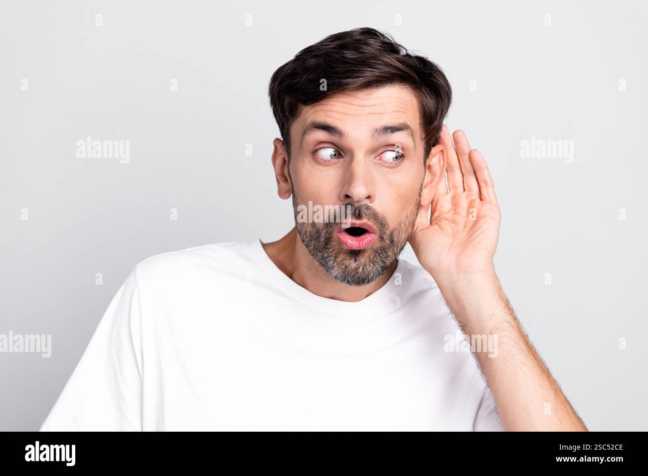 Junger bärtiger Mann, der mit neugierigem Ausdruck vor leerem Hintergrund genau zuhört, in lässigem weißem Outfit Stockfoto