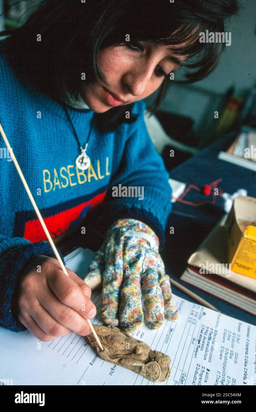 Eine Frau reinigt ein kleines Artefakt vorsichtig mit einem Stock, während sie einen blauen Pullover trägt, auf dem „BASEBALL“ steht. Stockfoto