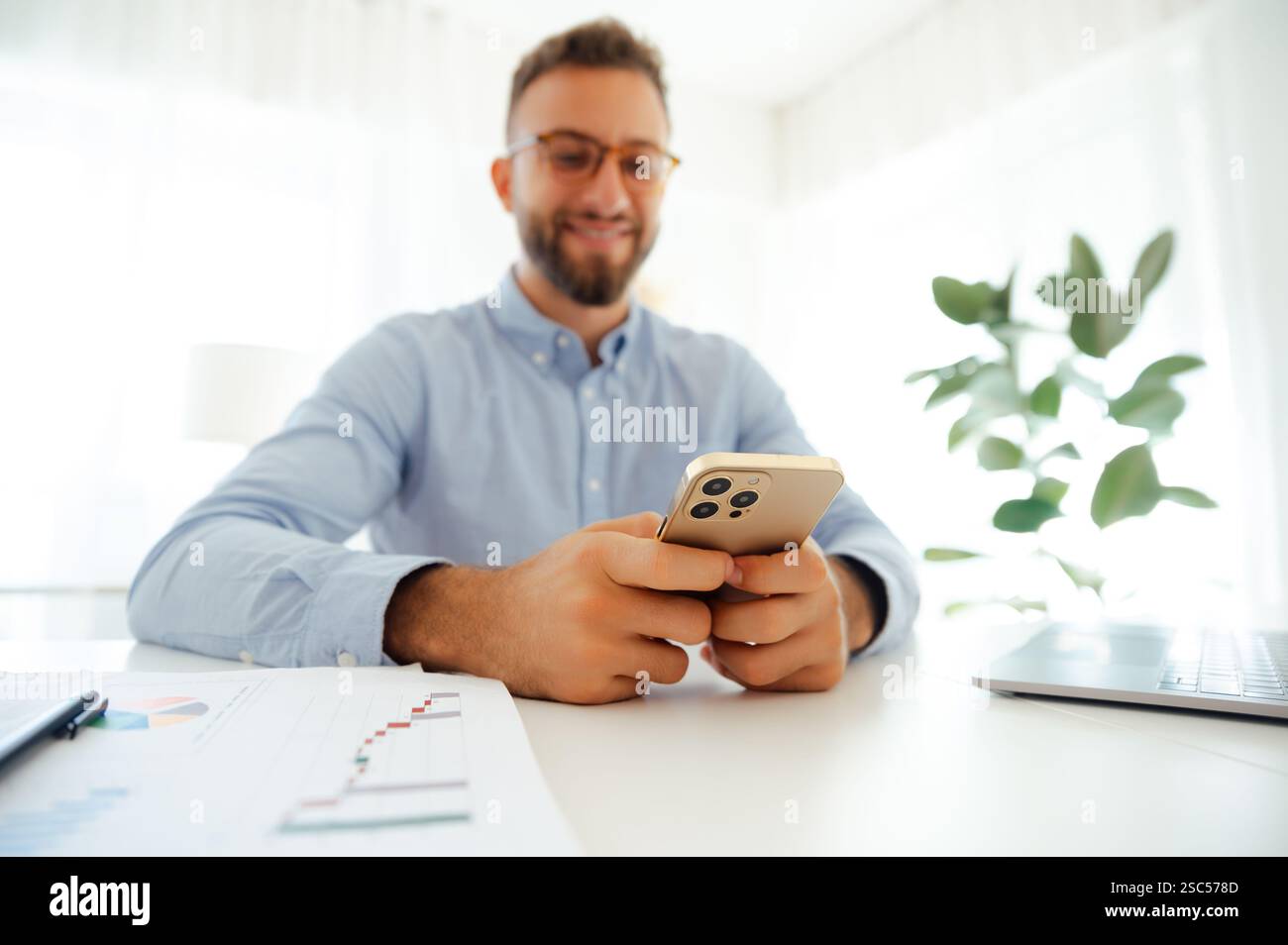 Unfokussierter Geschäftsmann mit Brille in einem blauen Hemd, der ein Smartphone benutzt, während er an einem Schreibtisch in einem hellen, modernen Büro sitzt. Konzept von Kommunikation, Fernarbeit und Online-Geschäft Stockfoto