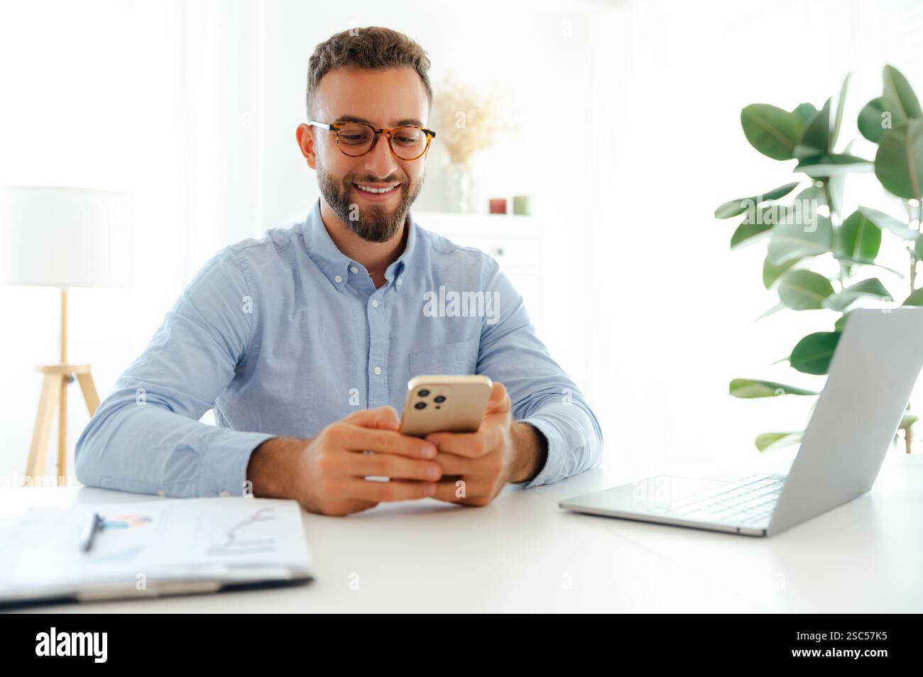 Lächelnder Geschäftsmann aus weißen oder arabien mit einer Brille in einem blauen Hemd, mit einem Smartphone, während er an einem Schreibtisch in einem hellen, modernen Büro sitzt. Konzept von Kommunikation, Fernarbeit und Online-Geschäft. Stockfoto
