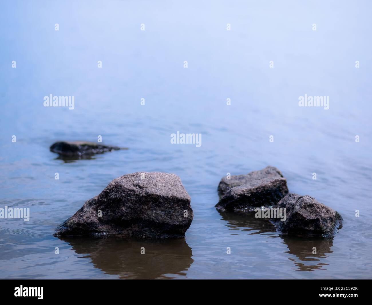 Minimalistische Zusammensetzung von Felsen, die aus ruhigem Wasser auftauchen, schaffen eine friedliche und ruhige Seeszene mit einem weichen blauen Farbverlauf. Stockfoto