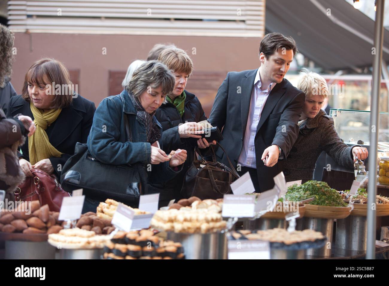 Leute, die an einem belebten Marktstand in London einkaufen und verschiedene Backwaren und Salate untersuchen. Stockfoto Leute, die an einem belebten Marktstand in London einkaufen und verschiedene Backwaren und Salate untersuchen. Stockfoto