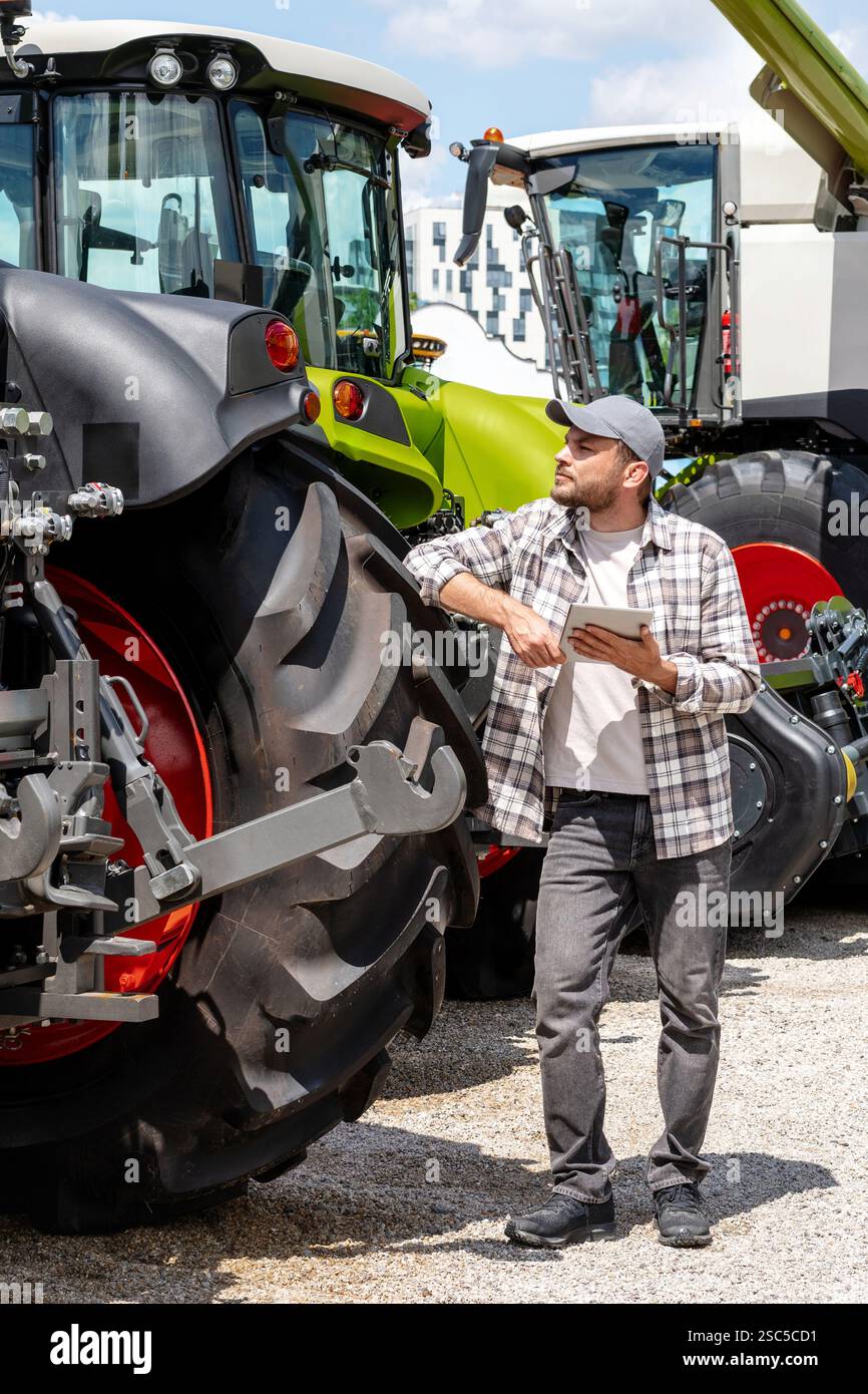 Händler oder Lieferant von Landmaschinen. Kaukasischer Mann in kariertem Hemd steht neben dem Traktor und benutzt ein digitales Tablet. Stockfoto