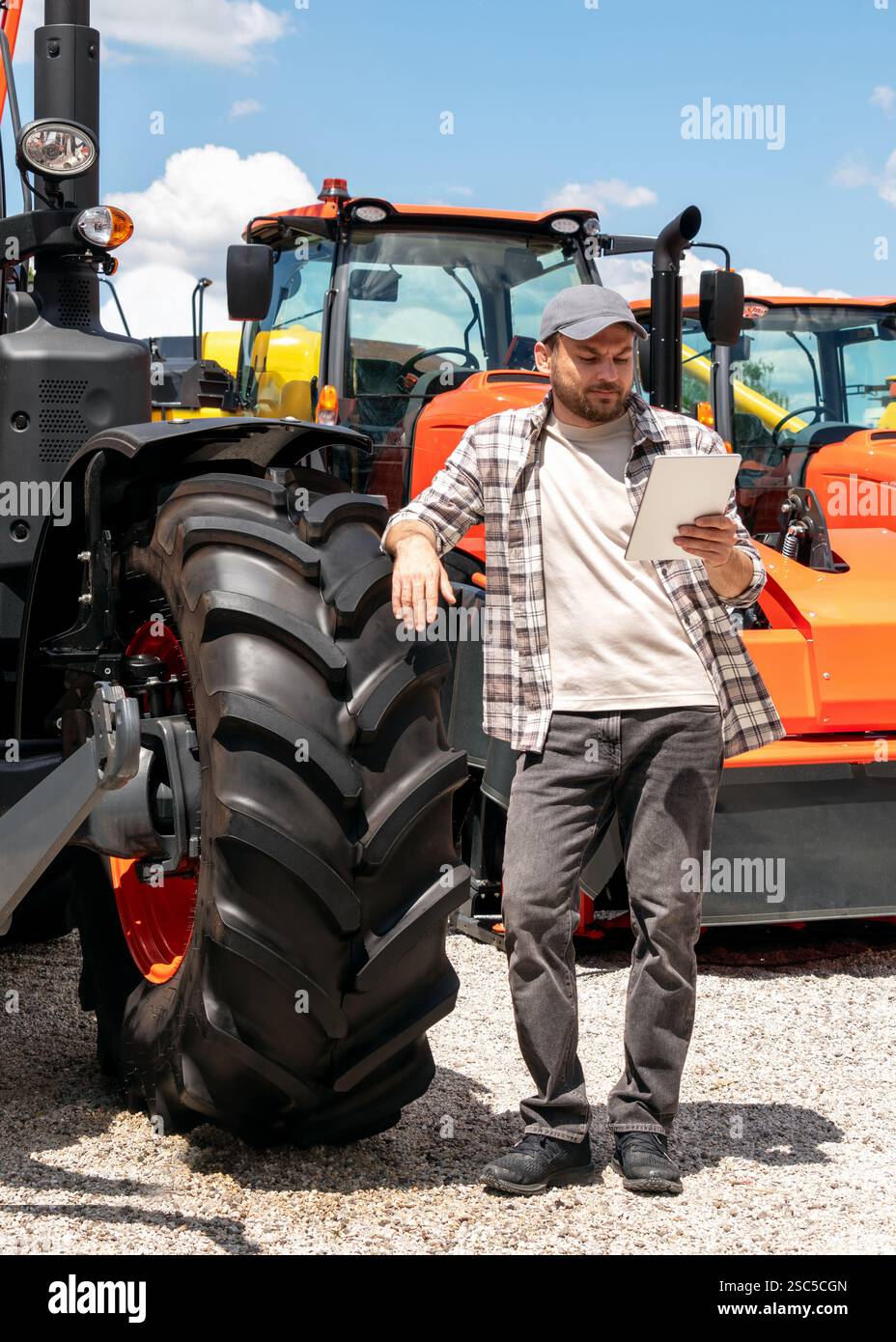 Händler oder Lieferant von Landmaschinen. Kaukasischer Mann in kariertem Hemd steht neben dem Traktor und benutzt ein digitales Tablet. Stockfoto