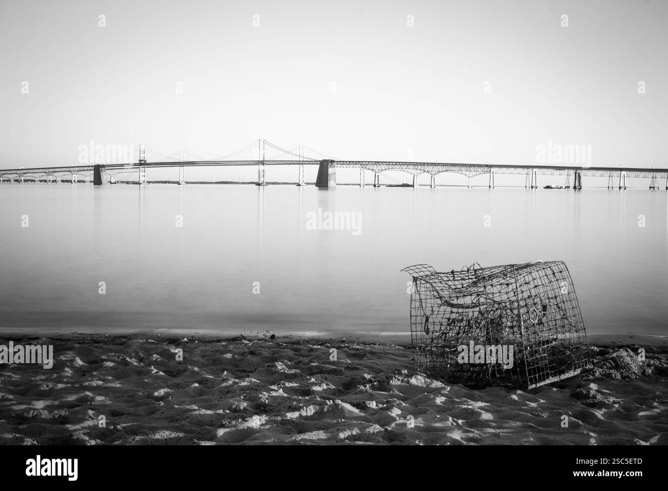 Ein Graustufenbild der Chesapeake Bay Bridge, die das Wasser überspannt. Eine ausrangierte Krabbenfalle liegt an der Sandküste. Stockfoto