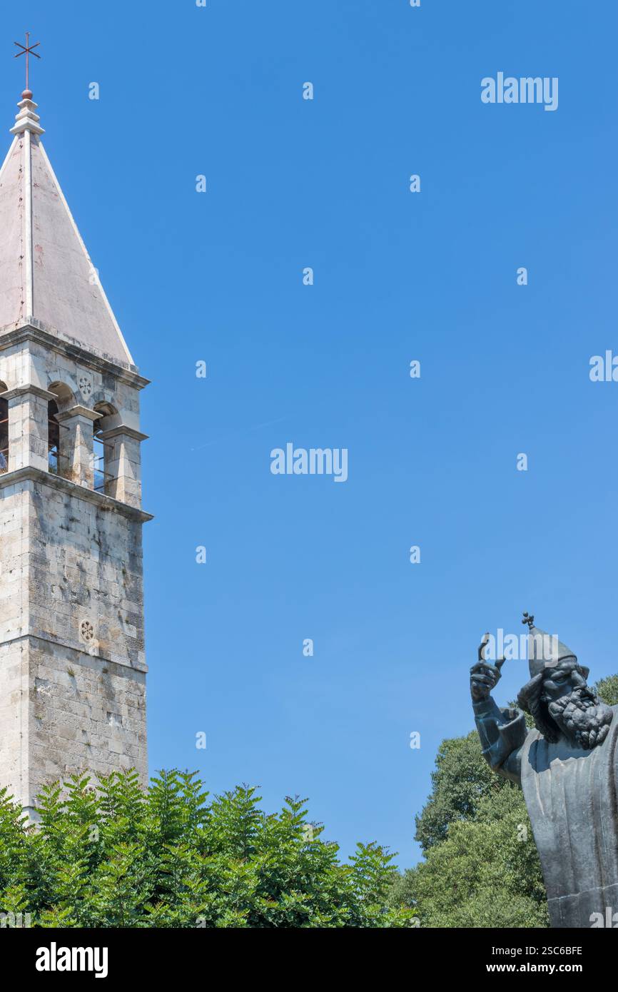Eine ruhige Szene mit einem historischen Glockenturm neben einer hohen Statue, umgeben von üppigem Grün, unter einem klaren blauen Himmel, der an Kultur und Geschichte erinnert Stockfoto