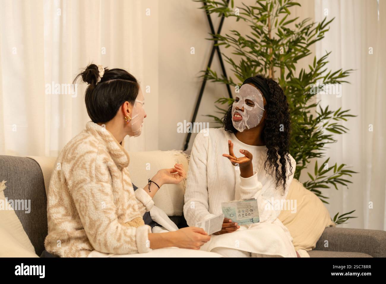 Zwei verschiedene Freundinnen diskutieren über Schönheitsprodukte, während sie Gesichtsmasken tragen und bequem auf einem Sofa in ihrem Wohnzimmer sitzen Stockfoto