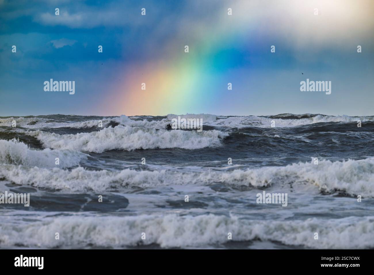 Ein lebhafter Regenbogen überzieht einen turbulenten Ozean mit stürzenden Wellen unter einem bewölkten Himmel. Stockfoto