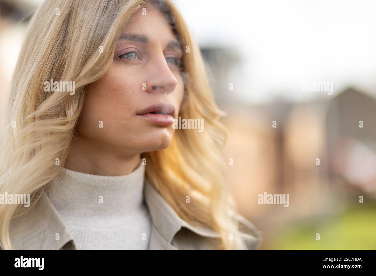 Eine Frau mit langen blonden Haaren steht draußen, im Sonnenlicht getaucht und blickt nachdenklich in die Ferne. Sie trägt einen leichten Pullover und einen selbstbewussten Ex Stockfoto