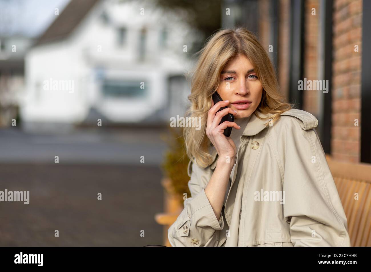 Eine junge Frau mit langen blonden Haaren sitzt auf einer Bank und trägt einen beigefarbenen Trenchcoat. Sie führt ein Telefongespräch, während sie einen sonnigen Tag in sich verbringt Stockfoto