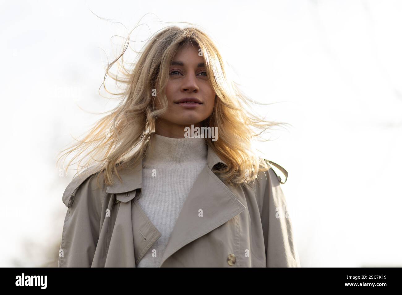 Eine Frau mit langen, welligen blonden Haaren steht draußen. Ihr Mantel wiegt sanft im Wind. Das sanfte Sonnenlicht scheint auf sie und schafft ein ruhiges atmos Stockfoto