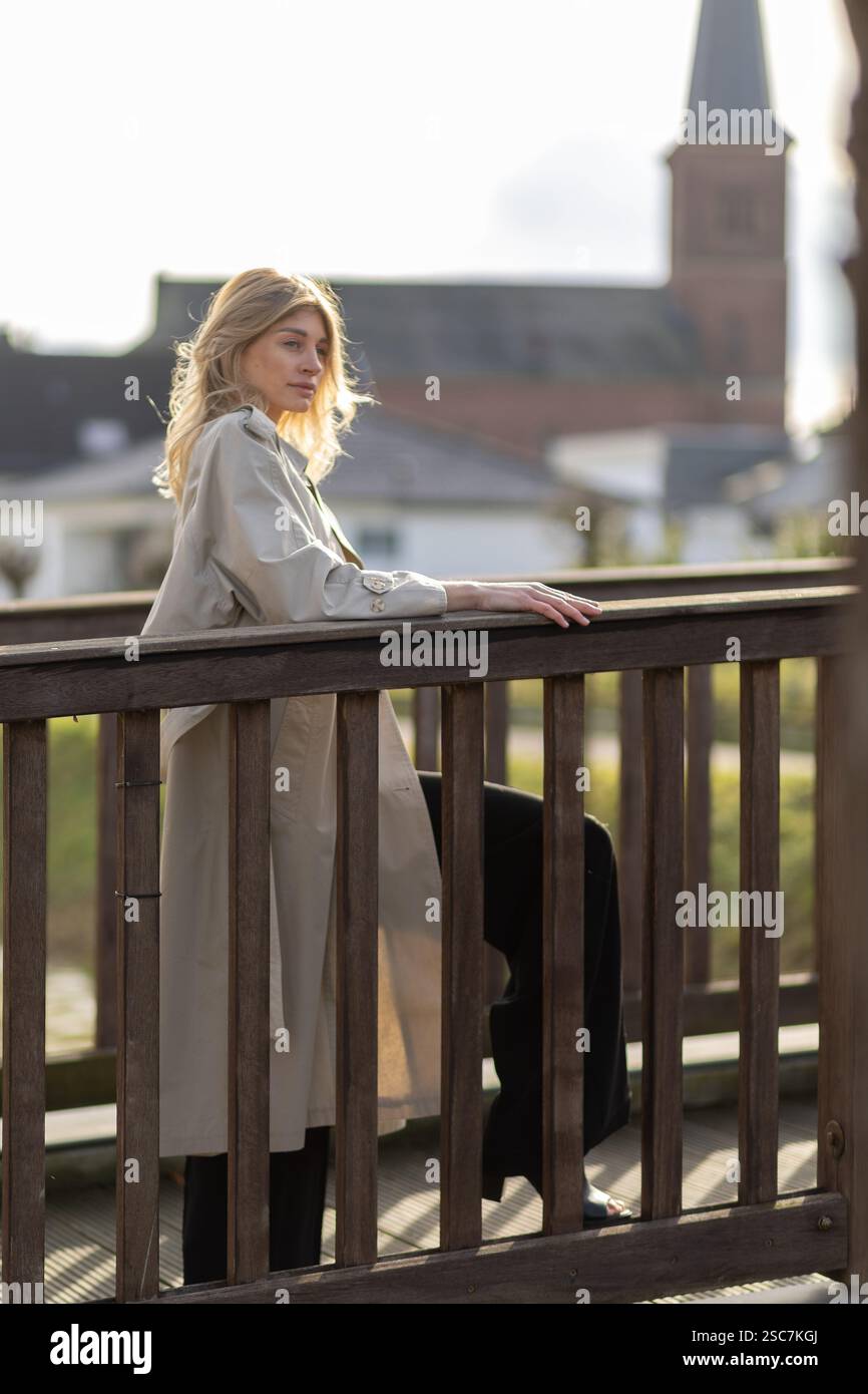 Eine Frau steht elegant auf einer Holzbrücke, stilvoll in einem Mantel gekleidet. Der Kirchturm ragt im Hintergrund, während sanftes Sonnenlicht ihren Ser hervorhebt Stockfoto