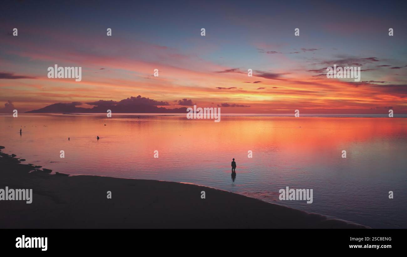 Atemberaubender Blick aus der Luft auf einen lebendigen Sonnenuntergang, der sich in ruhigen Gewässern des Ozeans spiegelt. Ein einsamer Mann steht an der Küste und schafft eine ruhige und malerische Szene in einem tropischen Paradies Stockfoto