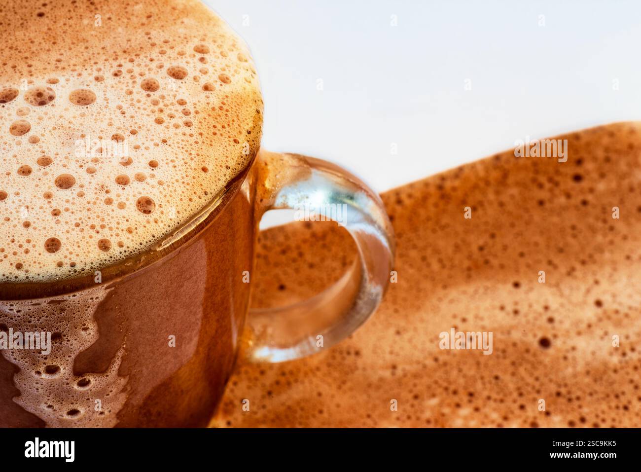 Teil eines heißen schäumenden Schokoladengetränks in Glasbecher. Getränk überläuft auf Becher und auf weißem Hintergrund, Nahaufnahme. Stockfoto