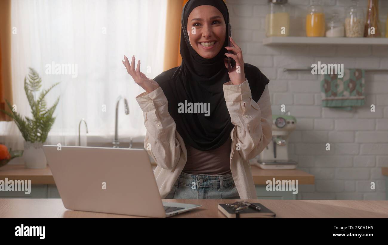 Glückliches lächelndes islamisches Mädchen muslimische Frau im Hijab Dame indische weibliche Hausfrau Klientin bestellen Essenslieferung in der Küche mit Laptop sprechendem Handy Stockfoto