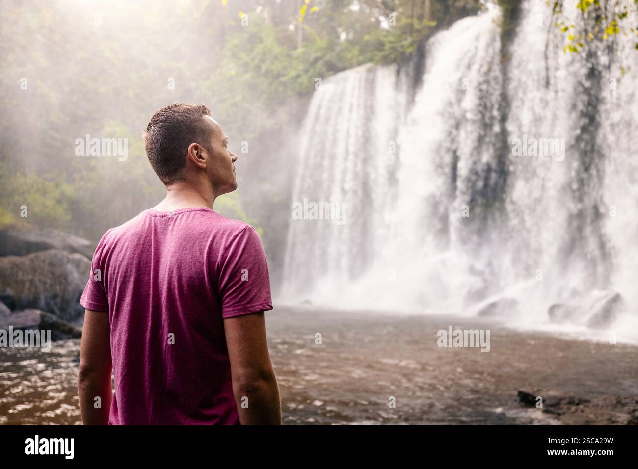 Mann vor hohem Wasserfall in Bergen in tropischer Landschaft. Reisende in Kamobodscha. Stockfoto