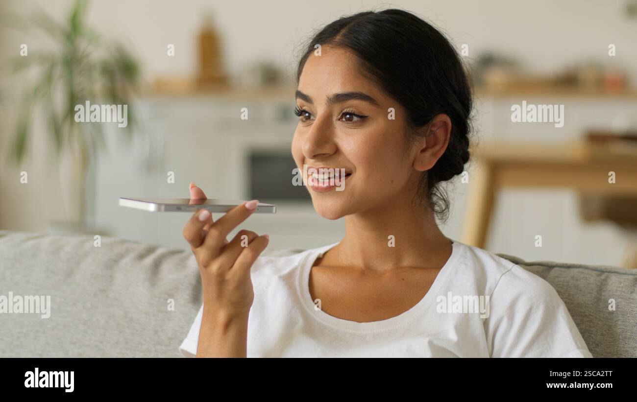Glückliche lächelnde indische Frau zu Hause Arabische positive arabische Mädchen weibliche Dame im Wohnzimmer Handy sprechendes Mikrofon Smartphone Mikrofon Mikrofon sprechen Stockfoto