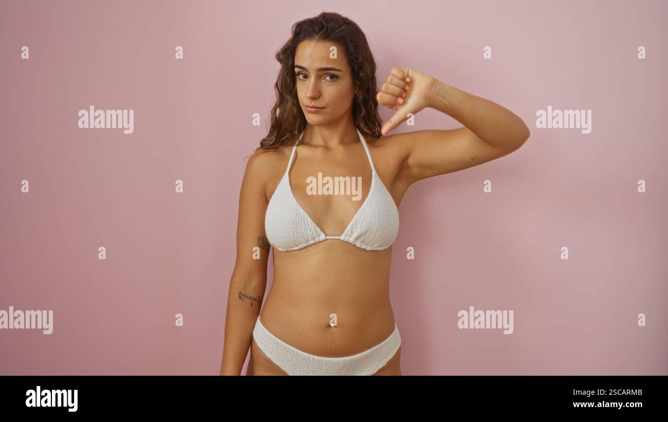 Eine junge Frau in einem weißen Bikini, die die Daumen nach unten über einen isolierten rosa Wandhintergrund hält Stockfoto