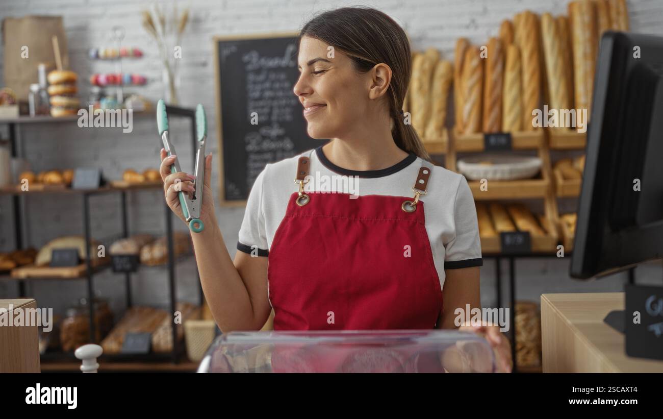 Die junge Frau in einer roten Schürze arbeitet in einer Bäckerei mit einer Küchenzange, umgeben von Regalen mit frischem Brot und Gebäck Stockfoto