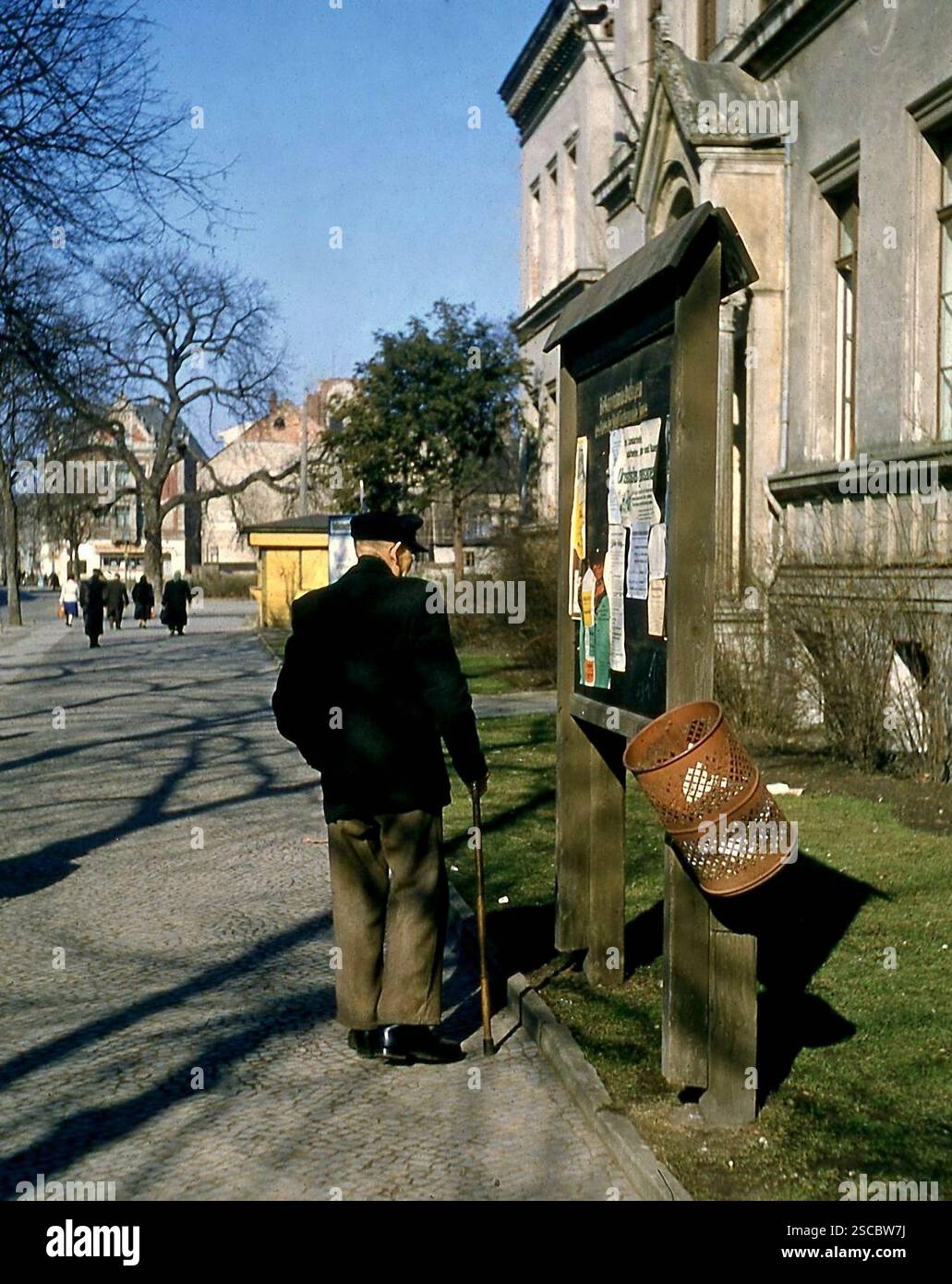 Ein alter Mann mit Mütze und Rohrstock liest Ansagen auf einer Tafel vor der Grundschule in Fürstenwalde. [Automatisierte Übersetzung] Stockfoto