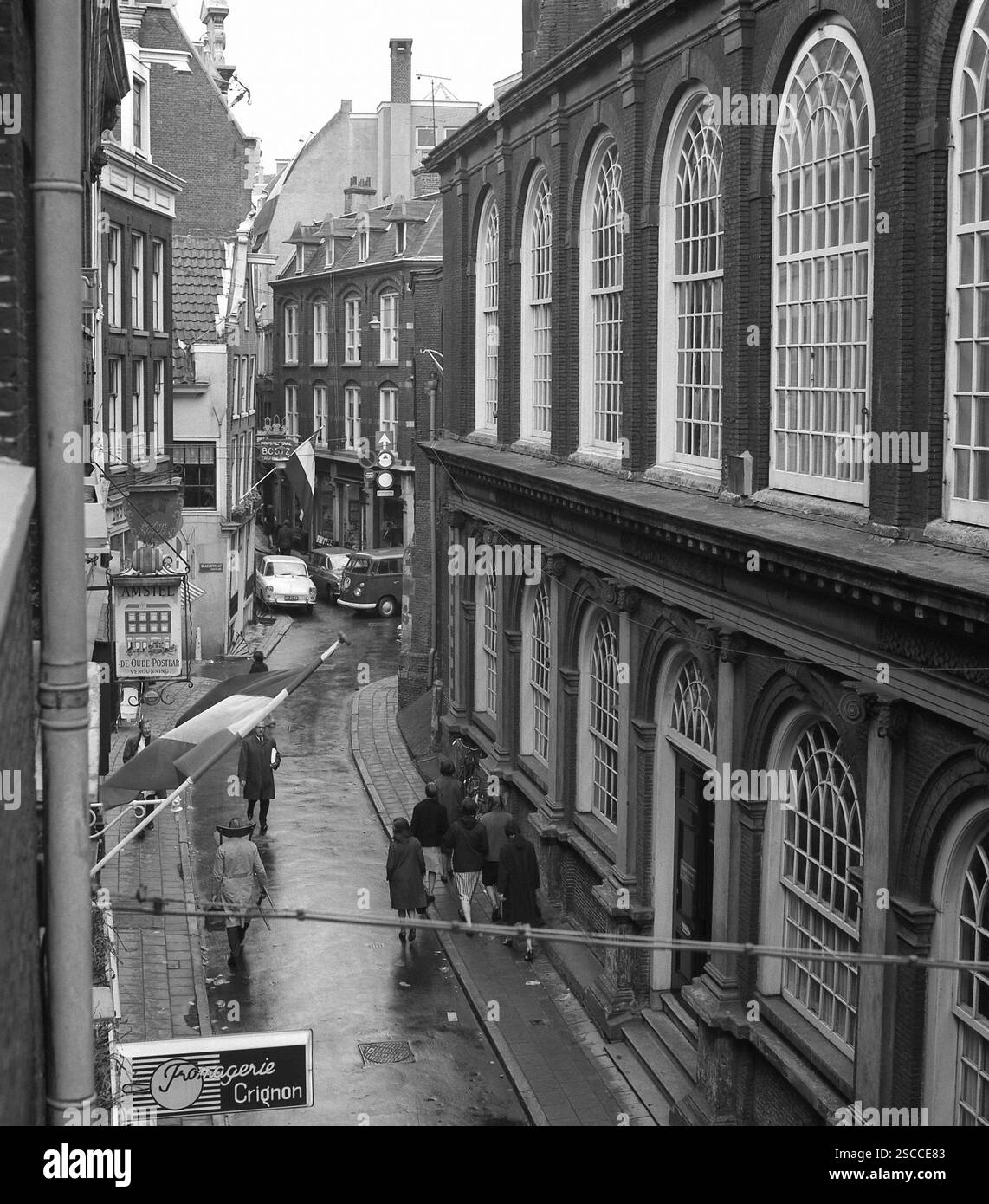Gasse in Amsterdam. Bild zeigt die Passanten (vorne ein Mann mit Stuhl um den Hals), Flagge, Schilder (Fromagerie Crignon, Amstel, De Oude Postbar) und auf der Rückseite Autos (VW-Bus). Stockfoto