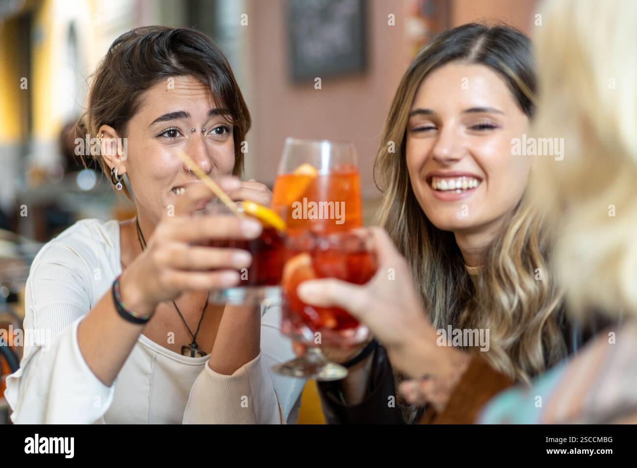 Drei junge Frauen, die mit Aperol Spritz und Cocktails in einem Café anstoßen, Freundschaft feiern und einen lebendigen sozialen Moment genießen. Stockfoto