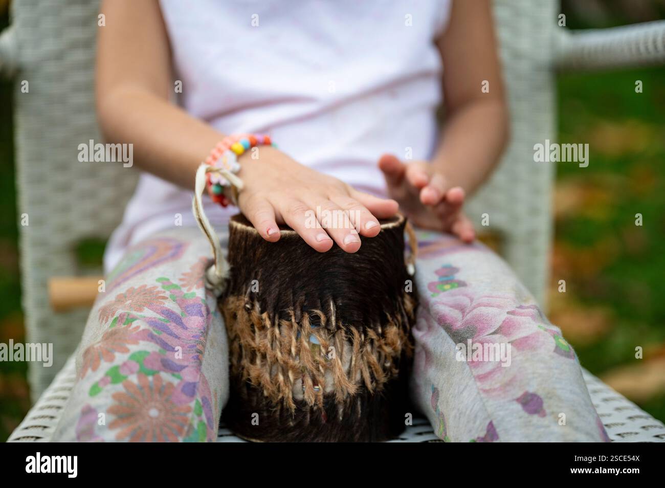 Ein Kind mit bunten Blumenhosen und perlenbesetzten Armbändern spielt eine kleine Handtrommel mit einem pelzbedeckten Körper, der auf einem weißen Korbstuhl sitzt Stockfoto