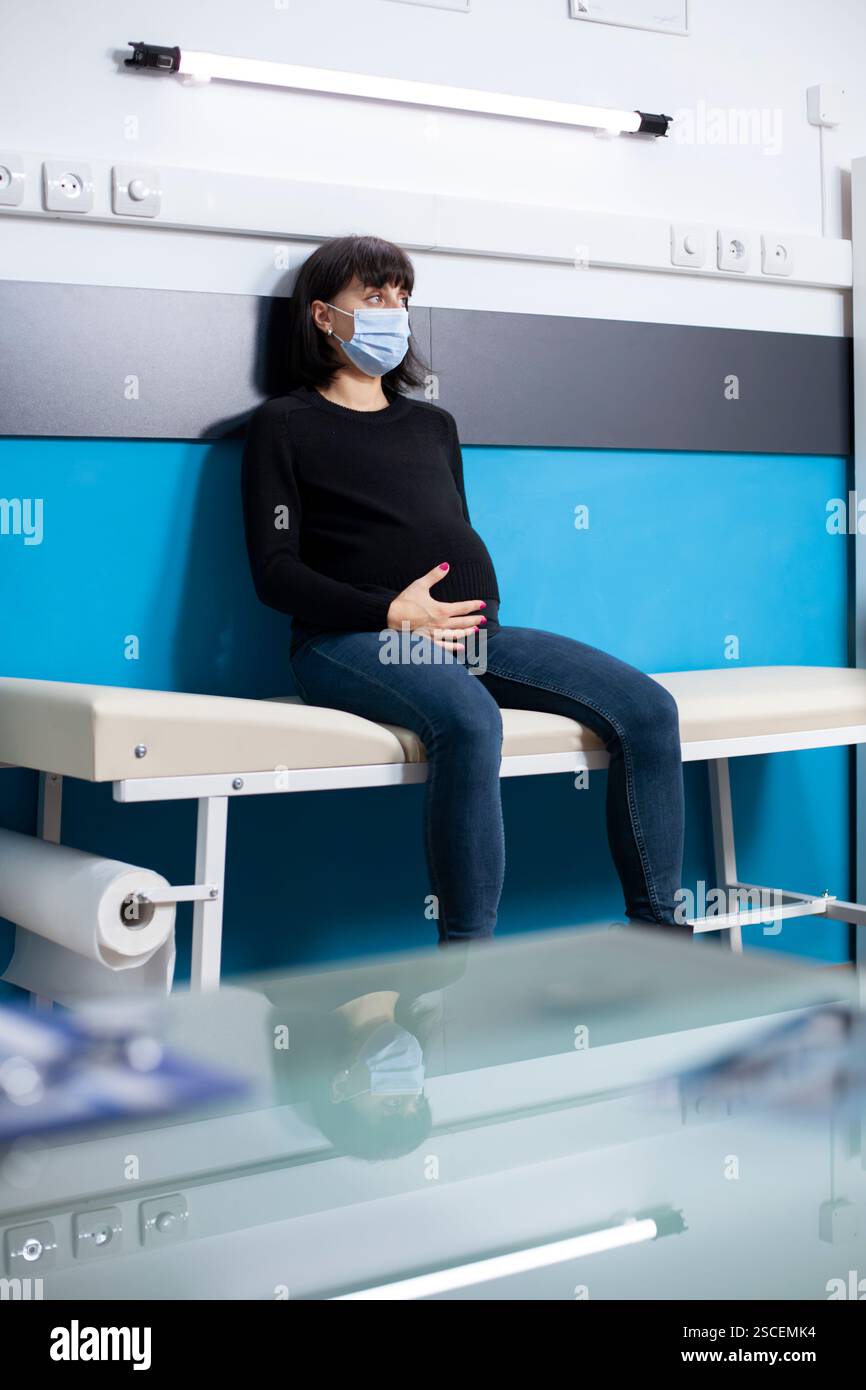 Weiße schwangere Frau in lässiger Kleidung sitzt ruhig auf dem Krankenhausbett und berührt sanft ihren Bauch. Junge werdende Mutter mit Gesichtsmaske, die auf die Mutterschaftsberatung in der Arztpraxis wartet. Stockfoto