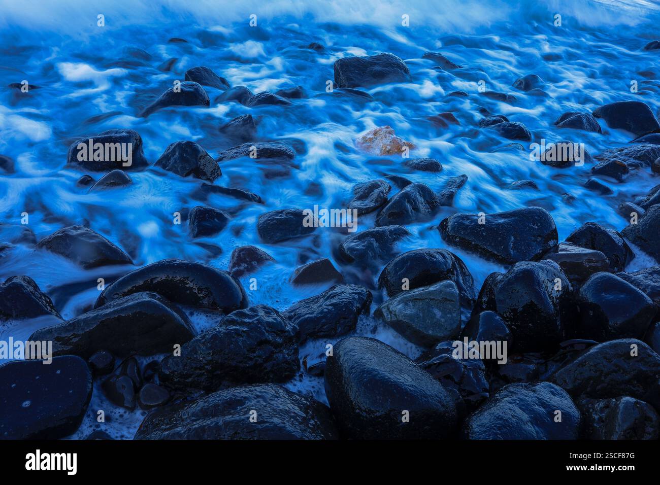 Lange ausgesetzte schwarze Vulkangesteine, darunter ein heller Stein, während nebelige Ozeanwellen über sich ziehen. Stockfoto
