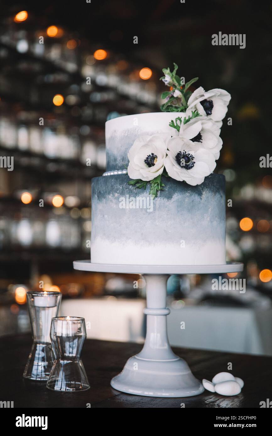 Ein wunderschön gefertigter, mehrstufiger Kuchen mit Farbverlauf und zarten weißen Blüten auf der Oberseite. Der Kuchen wird auf einem Stand in einem gemütlichen Ambiente präsentiert Stockfoto