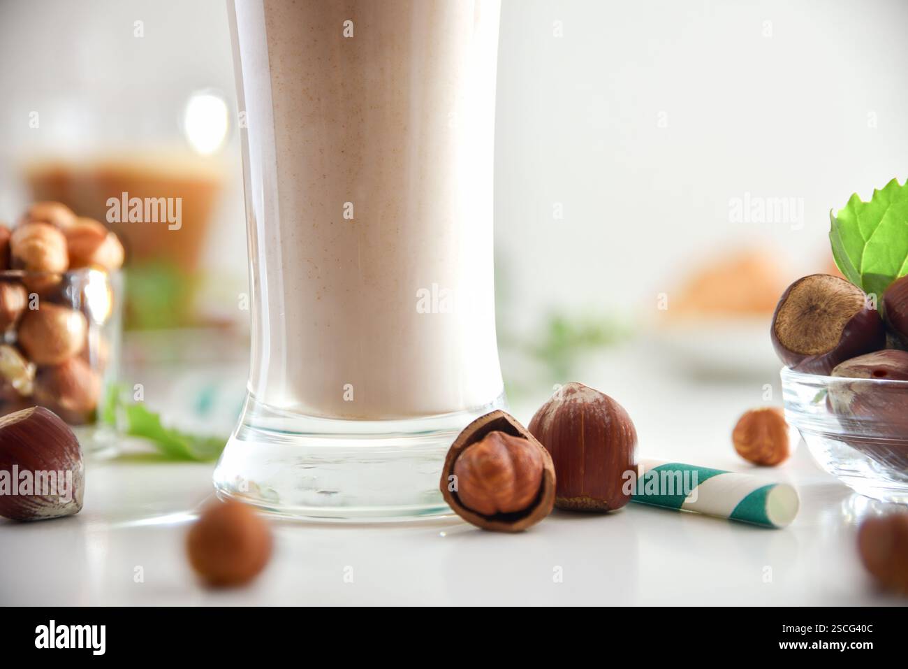 Detail aus Glas mit Haselnussgetränk mit Nüssen und Schüsseln voller Nüsse auf weißem Tisch und grünem Stroh mit isoliertem Hintergrund. Vorderansicht. Stockfoto