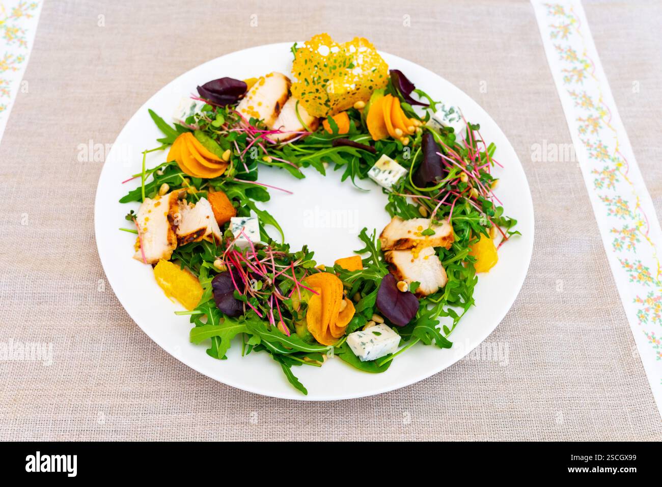 Farbenfroher Salat mit gegrilltem Huhn, Rucola und verschiedenen Gemüsesorten, die in kreisförmigen Mustern auf einem weißen Teller angeordnet sind. Die Einstellung ist Casua Stockfoto