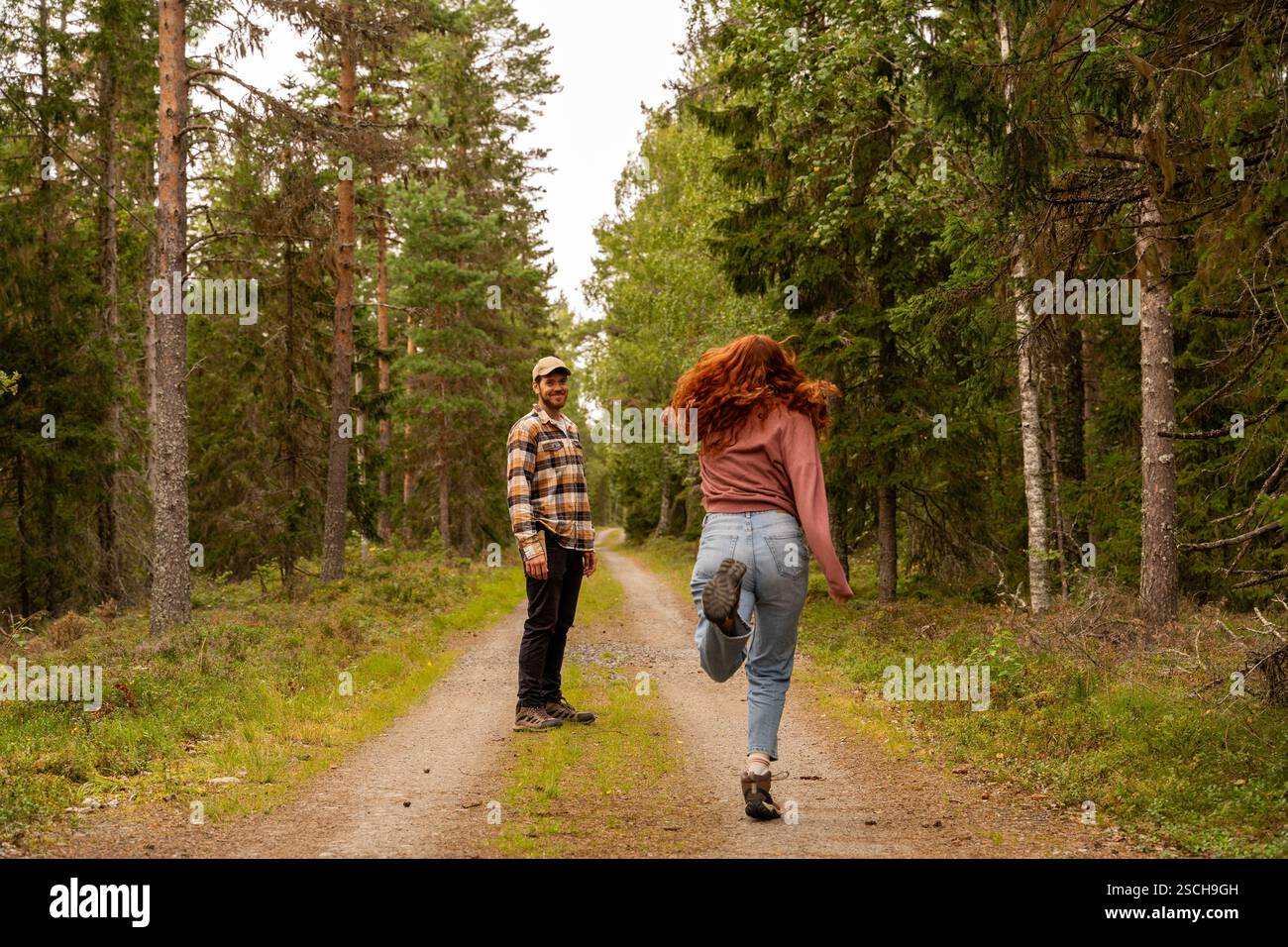 Ein Paar genießt einen gemütlichen Spaziergang auf einem unbefestigten Pfad in einem schwedischen Wald. Der Mann lächelt, während die Frau fröhlich überspringt, umgeben von üppigem Grün und Stockfoto
