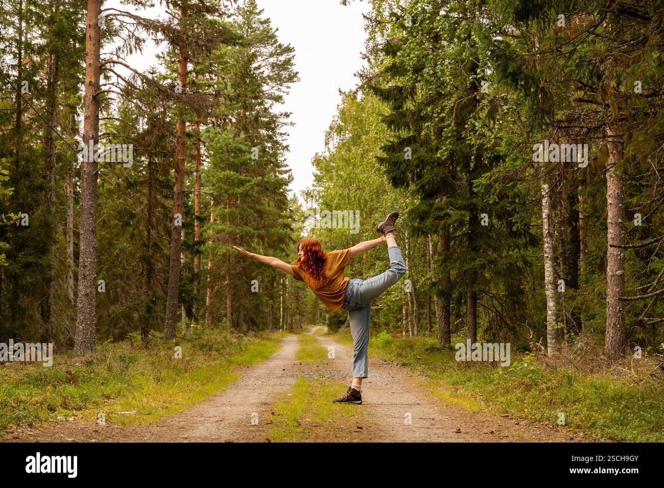 Eine Frau spielt eine Yoga-Pose auf einem Waldweg, der von üppigen Bäumen gesäumt ist, in Schweden, die Gleichgewicht und Verbindung mit der Natur verkörpert. Die heitere Atmosphäre Stockfoto