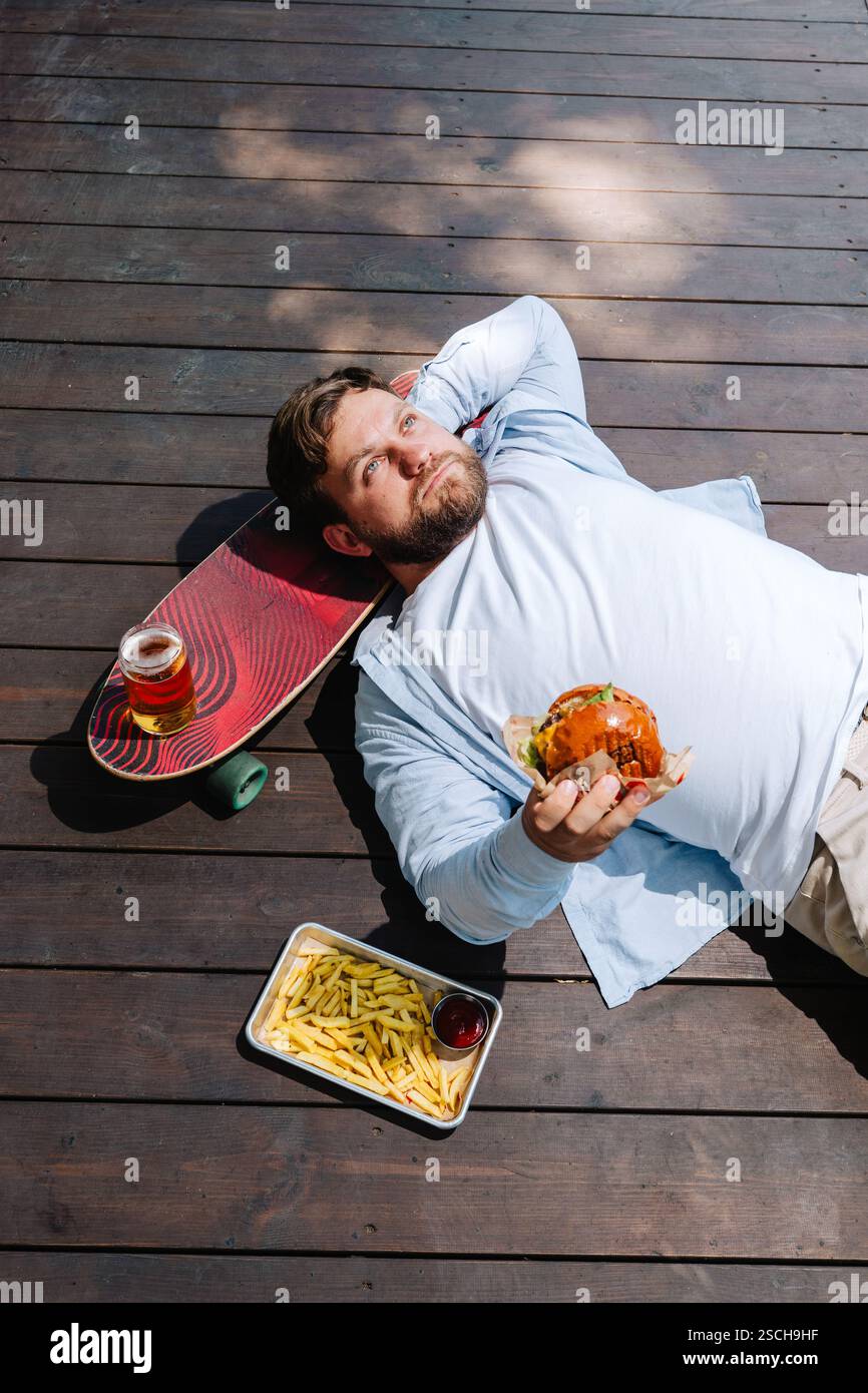 Eine Person, die einen entspannenden Moment auf einer Holzterrasse genießt und einen Burger hält. In der Nähe gibt es ein Skateboard, Pommes und ein Getränk, das die Essenz eines l einfängt Stockfoto