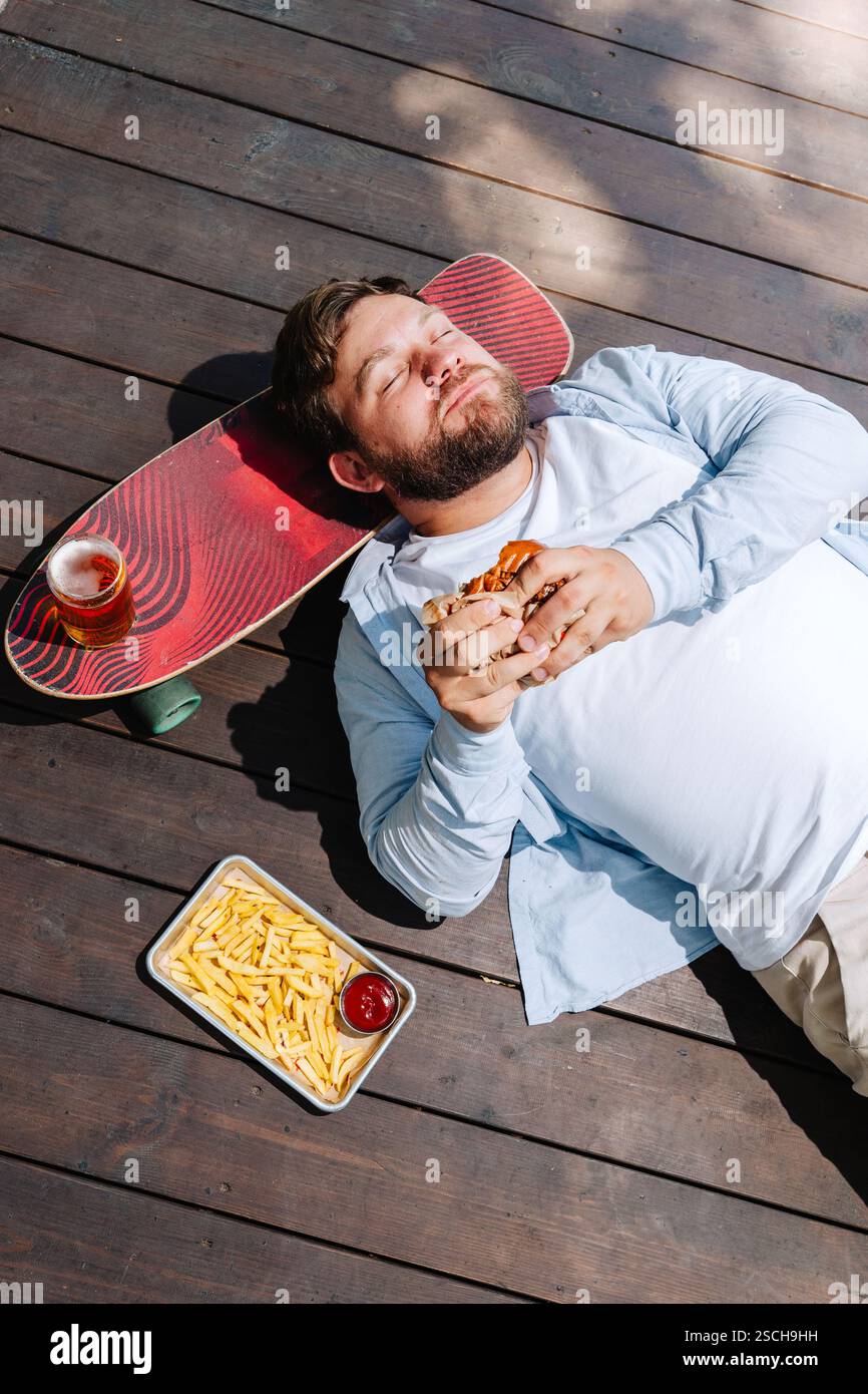 Ein Mann liegt auf einer Holzterrasse und genießt einen Burger mit einem Tablett Pommes Frites und einem Drink auf einem Skateboard neben ihm. Die Szene fängt ein entspanntes, draußen Stockfoto