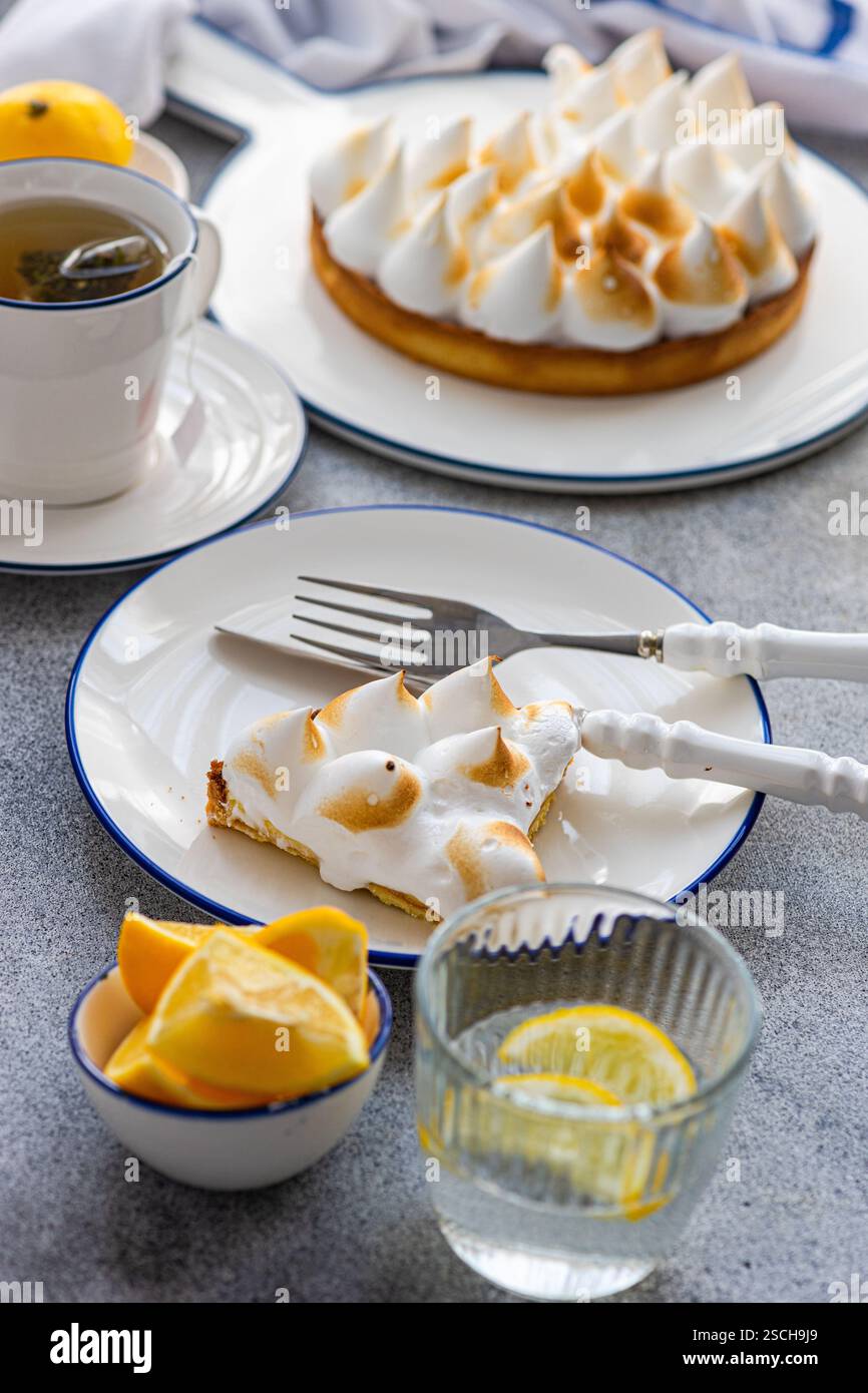 Ein Stück hausgemachter Zitronenkuchen mit goldenen Gipfeln, serviert auf einem Teller, begleitet von einer Tasse Tee, frischen Zitronenscheiben und einer Zitronengarnitur in einem Glas. Stockfoto