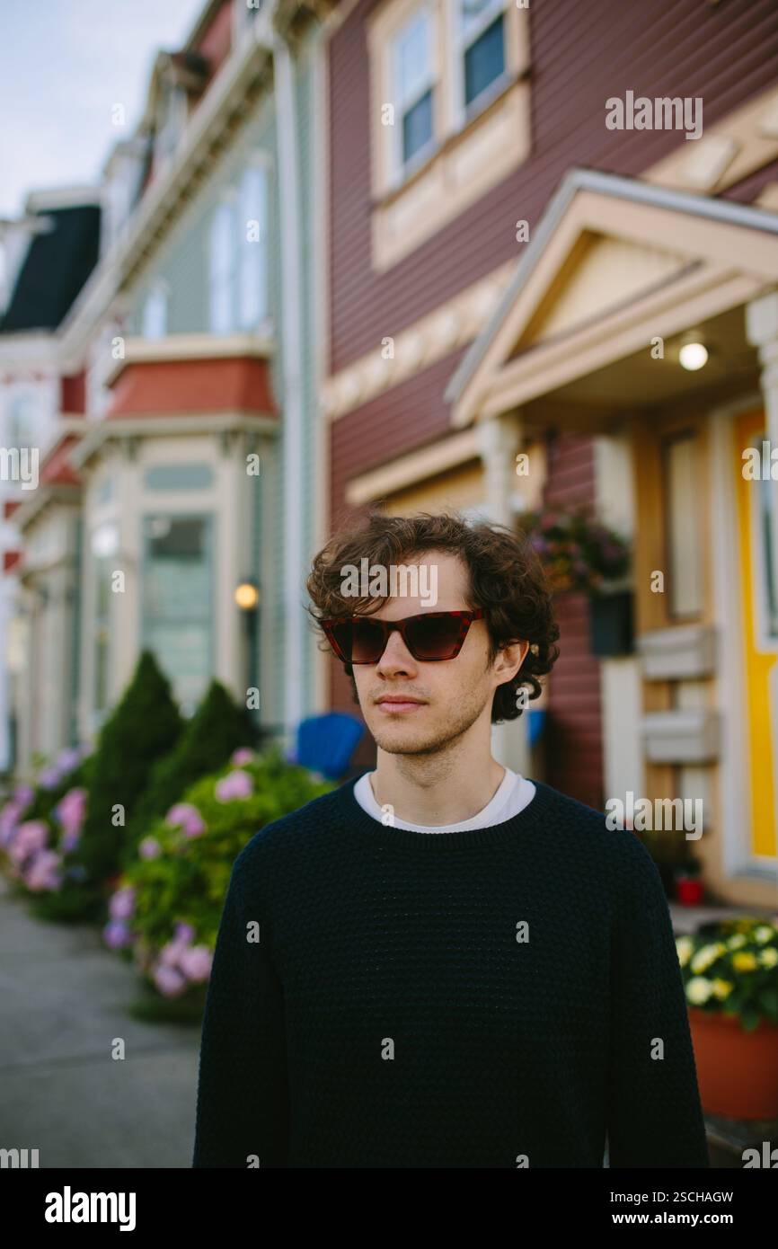 Ein stilvoller junger Mann steht selbstbewusst auf einer Straße in St. John's, trägt eine Sonnenbrille und einen lässigen Pullover. Die bunten Reihenhäuser im Hintergrund A Stockfoto