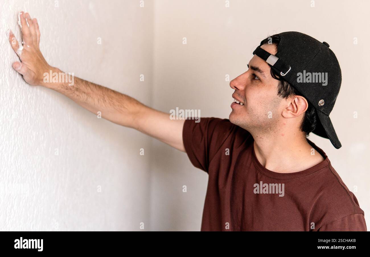 Ein junger Mann inspiziert eine Mauer während eines Renovierungsprojekts genau und ist bereit, seine nächste Aufgabe zu beginnen. Er trägt ein lässiges Outfit und eine Baseballmütze, er de Stockfoto