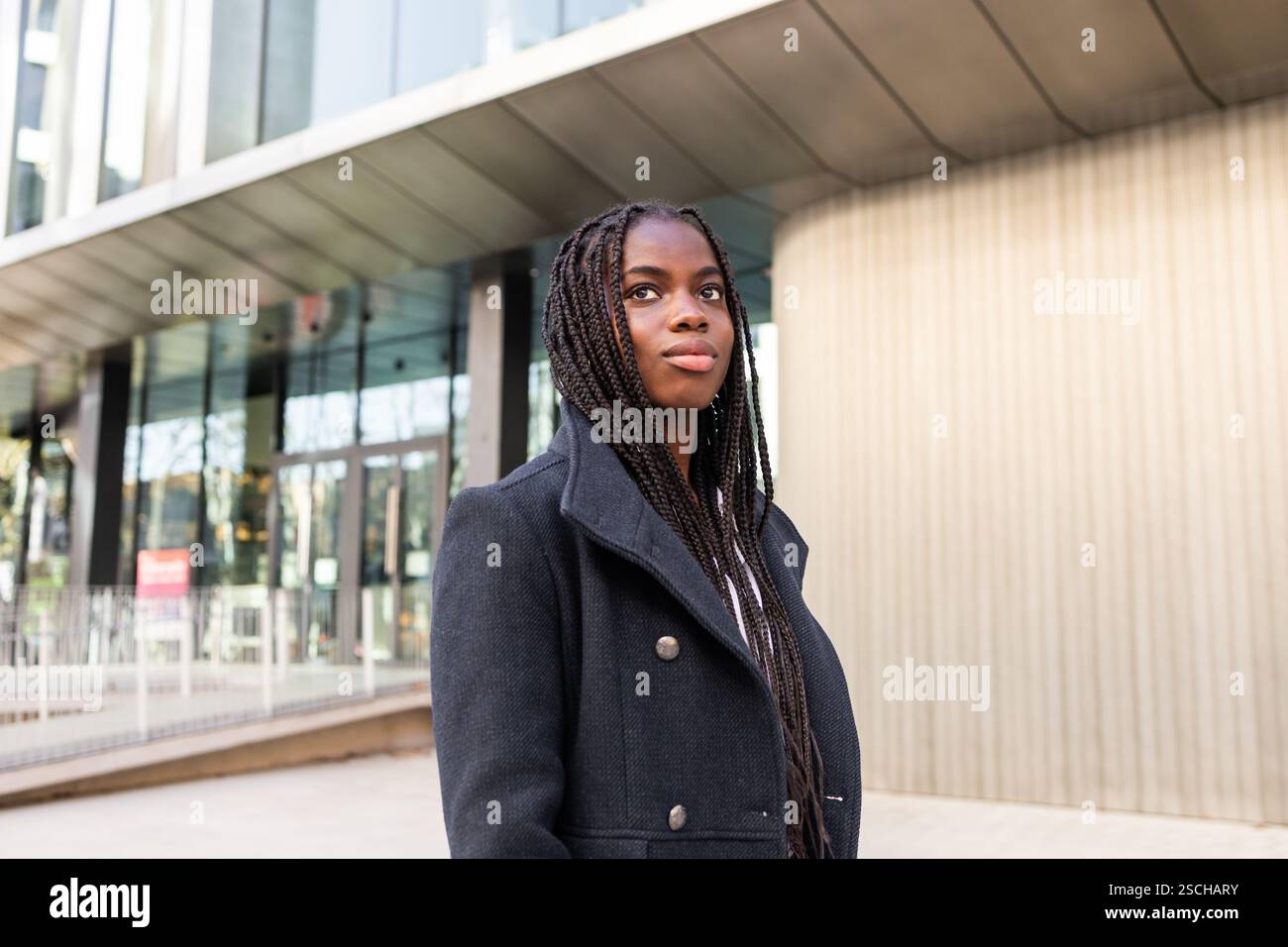 Eine junge, professionelle afroamerikanische Geschäftsfrau mit langen Zöpfen im Freien, die selbstbewusst in einem Geschäftsumfeld abblickt. Stockfoto