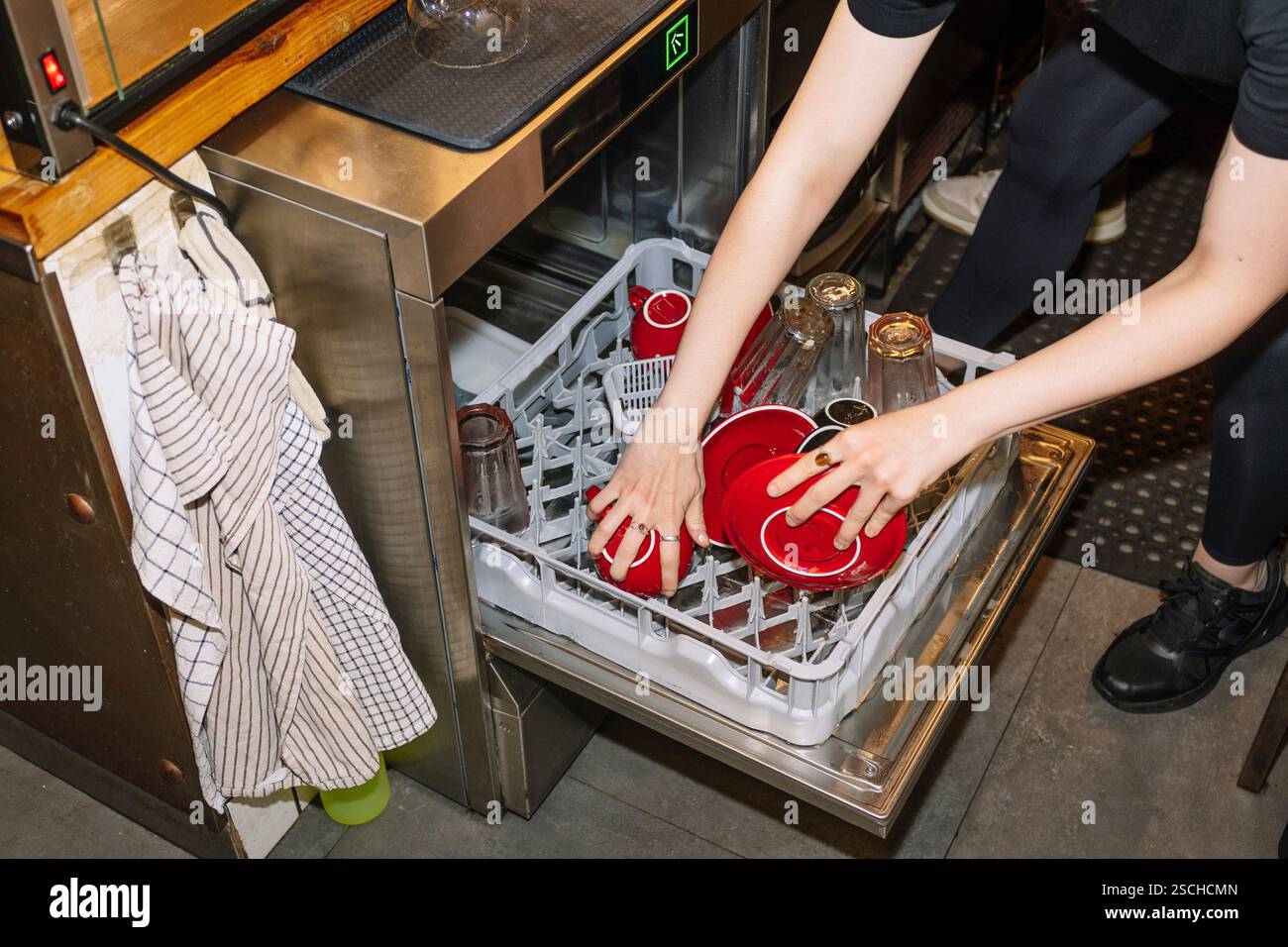 Eine Person lädt einen Geschirrspüler mit roten Kaffeetassen und Glasflaschen, um die Vorgänge hinter den Kulissen in einem speziellen Café oder Café envi zu erfassen Stockfoto