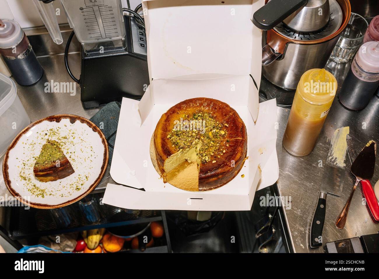Ein frisch gebackener Käsekuchen mit zerstoßenen Pistazien befindet sich in einer offenen Schachtel, in der Nähe befindet sich eine Scheibe. Die Szene zeigt eine Coffee-Shop-Küche mit Stockfoto