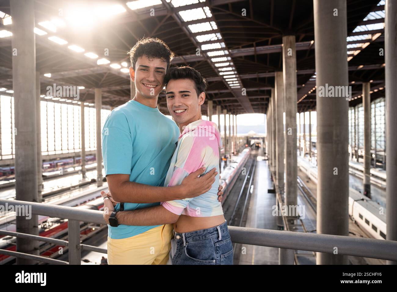 Ein fröhliches schwules Paar umarmt sich liebevoll an einem modernen Bahnhof, während Sonnenlicht durch die großen Fenster strömt, um einen Moment der Zuneigung und des Herzens festzuhalten Stockfoto