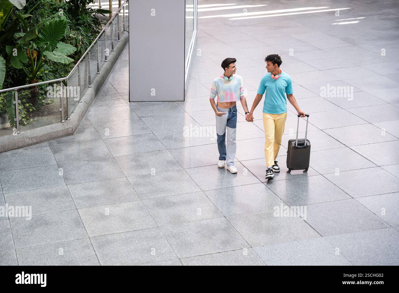 Ein fröhliches schwules Paar geht Hand in Hand durch ein Flughafenterminal mit Gepäck, genießt die Gesellschaft des anderen und sieht in lässiger Kleidung stilvoll aus. Stockfoto
