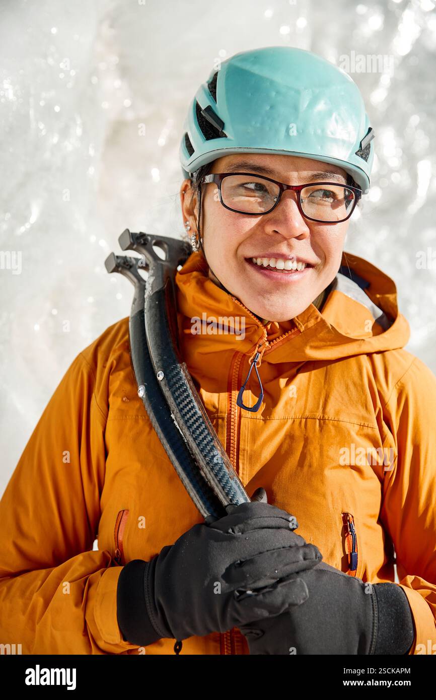 Junge asiatische Athletin in orange Jacke, blauem Helm und Axt Eiskletterausrüstung in der Nähe des gefrorenen Eiswasserfalls in den Bergen in der Nähe von Almaty City, Kasachs Stockfoto