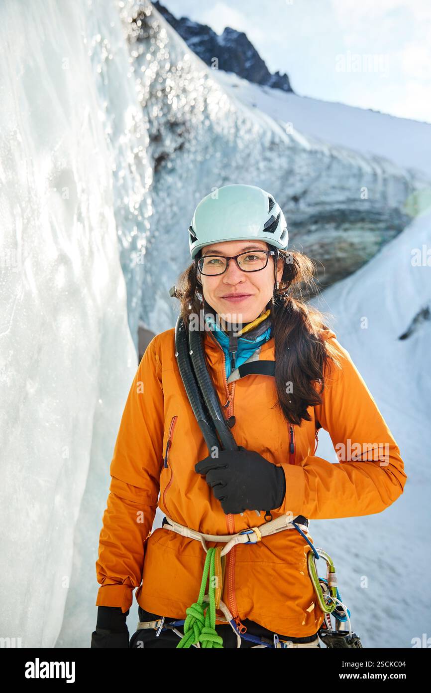 Junge asiatische Athletin in orange Jacke, blauem Helm und Axt Eiskletterausrüstung in der Nähe des gefrorenen Eiswasserfalls in den Bergen in der Nähe von Almaty City, Kasachs Stockfoto