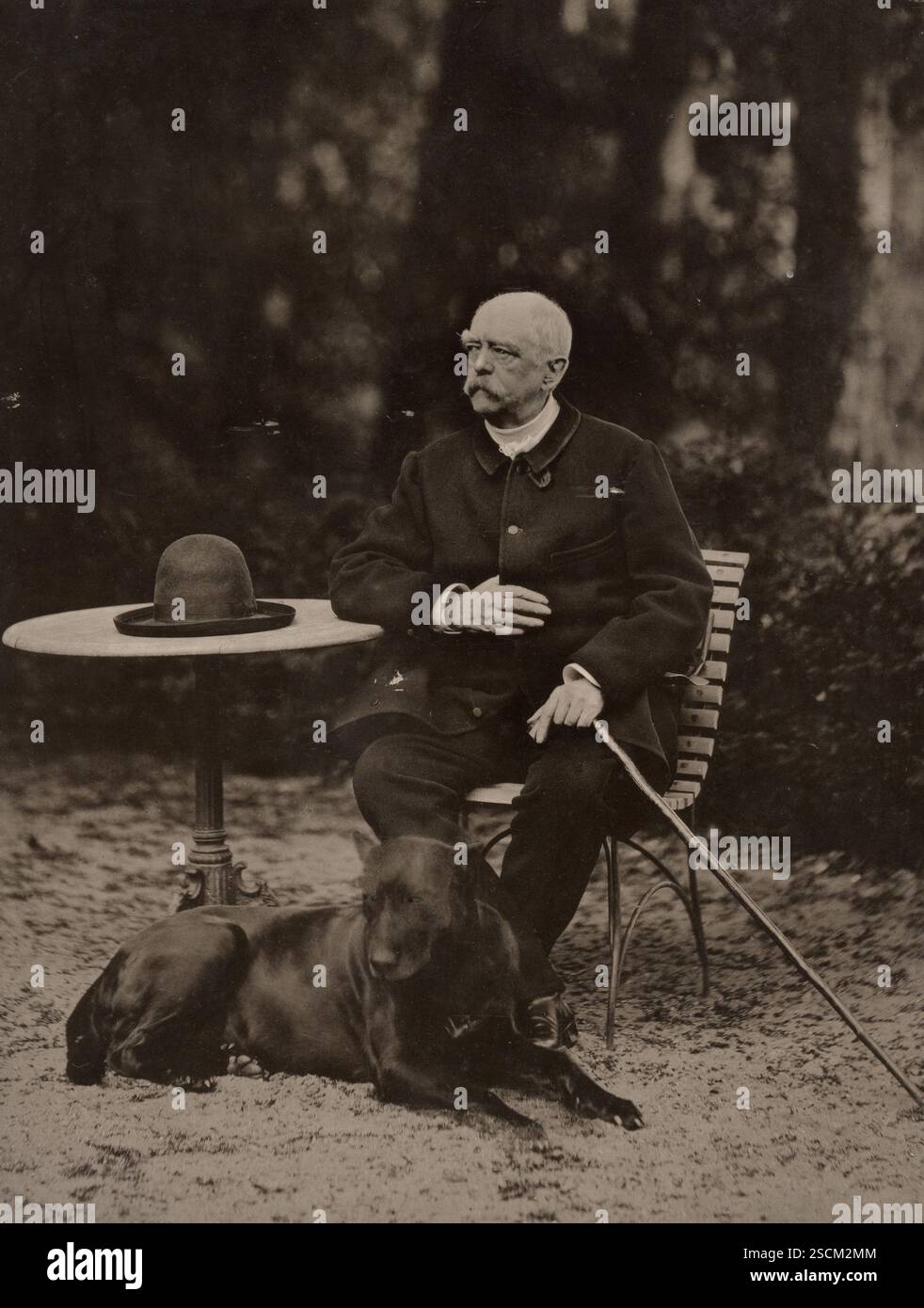 Bismarck, 1884. Porträt von Otto von Bismarck an einem Tisch draußen. Er hält einen Stock und seine Mütze liegt auf dem Tisch. Da ruht ein großer schwarzer Hund zu seinen Füßen. Stockfoto