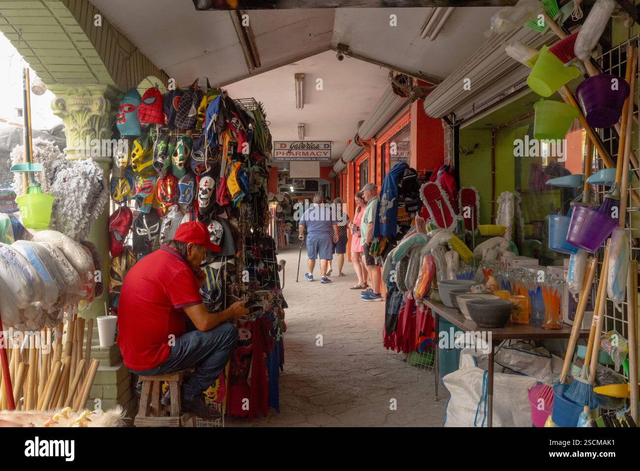 Verkäufer auf seinem Handy, umgeben von Gegenständen zum Verkauf, Masken, Mopps, Kleidung, Besen, Menschen auf dem Bürgersteig, in der Grenzstadt Nuevo Progreso, Mexiko. Stockfoto