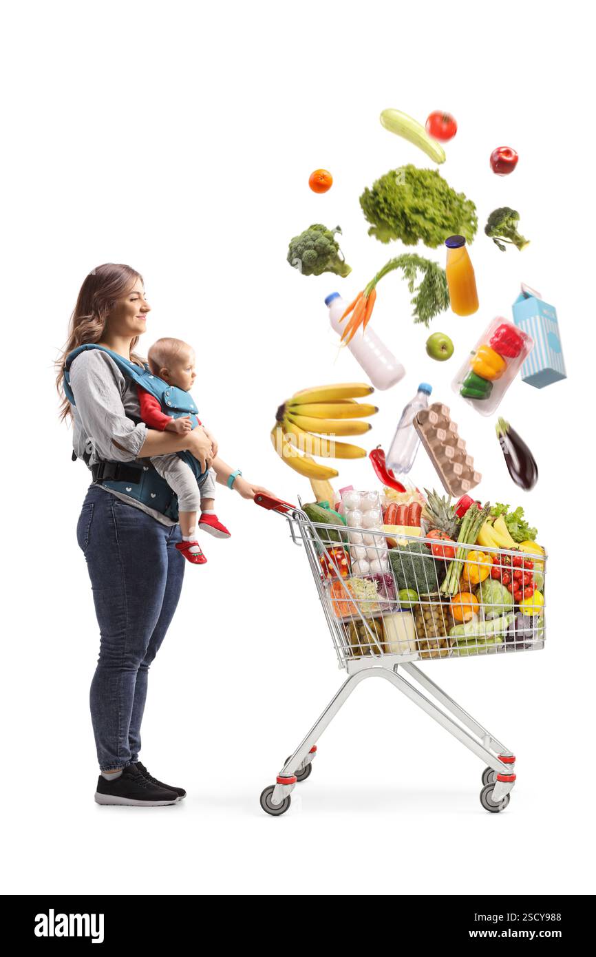 Mutter mit einem Baby in einer Tragetasche und einem Einkaufswagen voller herabfallender Lebensmittel, isoliert auf weißem Hintergrund Stockfoto