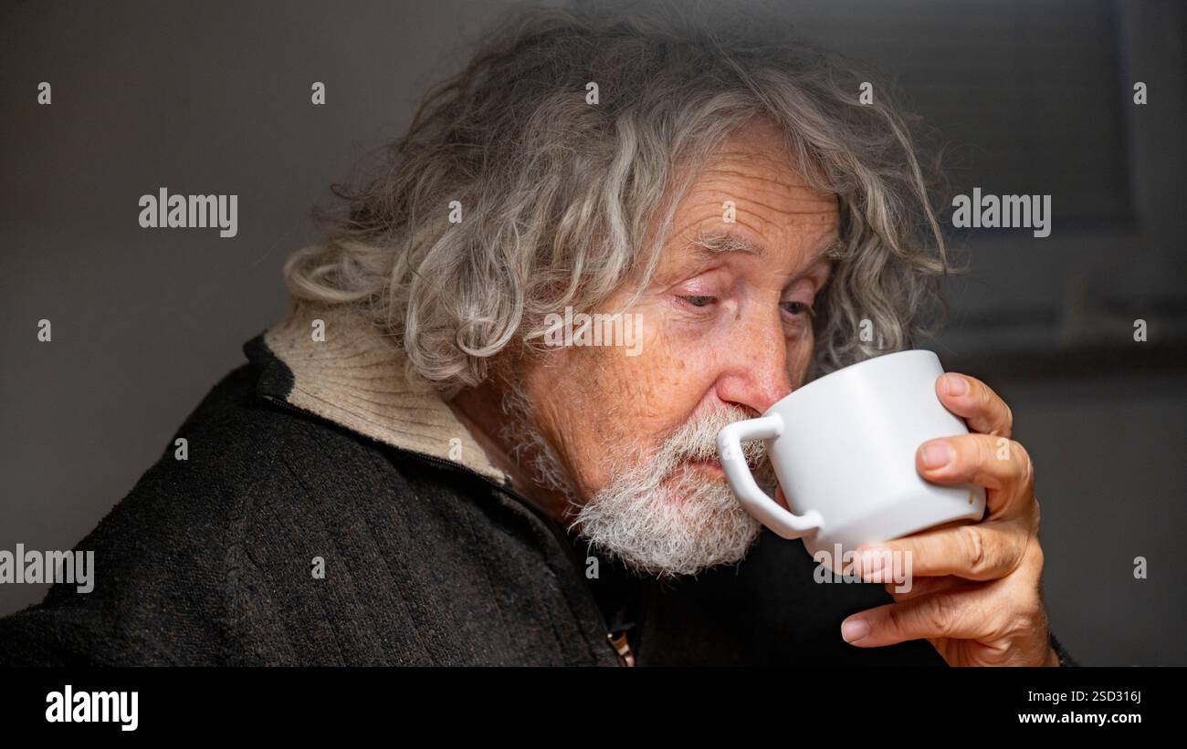 Ein älterer Mann mit lockigen grauen Haaren schlürft nachdenklich aus einem weißen Becher, sein Ausdruck ruhig und introspektiv. Stockfoto